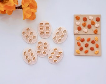 May include: A set of seven white plastic cookie cutters shaped like leaves, pumpkins, and acorns. The cutters are arranged on a white surface. Next to the cutters are two sheets of brown clay with cut-out shapes of leaves and pumpkins.