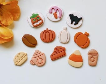 May include: A collection of 13 fall and Halloween themed clay ornaments. The ornaments are in various shapes and colors, including pumpkins, a coffee cup, a pie, a candy corn, a ghost, a black cat, and a jack-o-lantern.