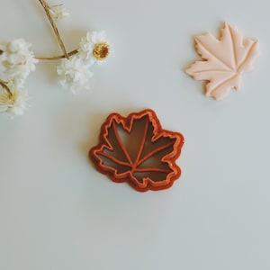 May include: A red plastic maple leaf cookie cutter. The cutter is on a white surface.