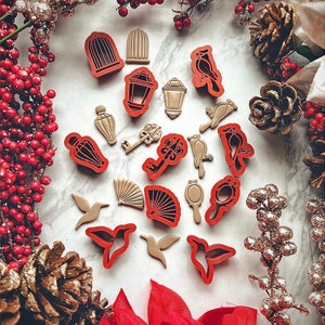 May include: A set of 16 red and gold cookie cutters featuring various shapes, including birds, keys, lanterns, and a fan. The cutters are arranged on a white surface with decorative pine cones and red berries.