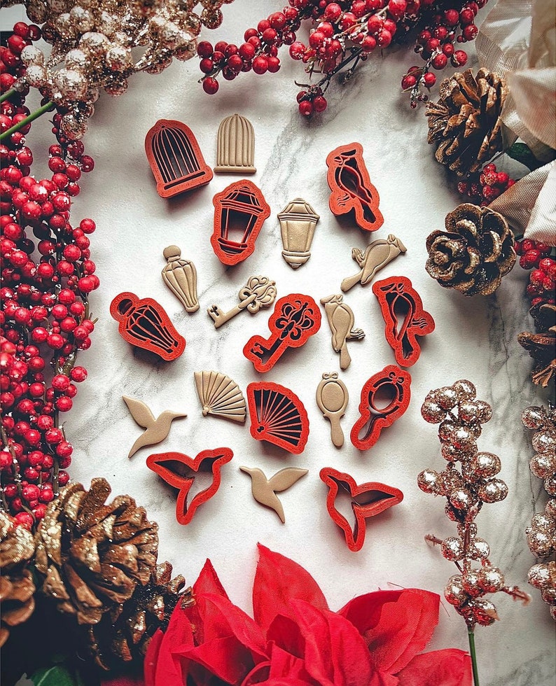 May include: A set of 16 red and gold cookie cutters featuring various shapes, including birds, keys, lanterns, and a fan. The cutters are arranged on a white surface with decorative pine cones and red berries.