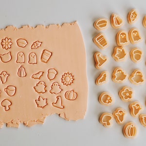 May include: A set of 20 cookie cutters in various fall and Halloween shapes, including pumpkins, ghosts, bats, leaves, acorns, and a coffee cup. The cutters are made of a light beige plastic and are arranged on a white surface.