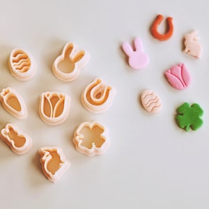May include: A set of 11 plastic cookie cutters in various shapes, including a bunny, a horseshoe, a clover, a carrot, a tulip, an egg, and a heart. The cutters are in shades of pink, green, orange, and brown.