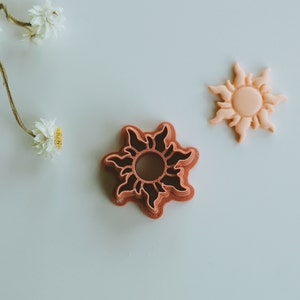 May include: Two sun-shaped cookie cutters, one in a light peach color and the other in a dark red color. The red cookie cutter is a detailed design with a raised center.
