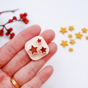 May include: A white plastic star-shaped cookie cutter with three different sized stars. The cutter is being held in a hand. There are several gold star-shaped cookies scattered on a white surface.