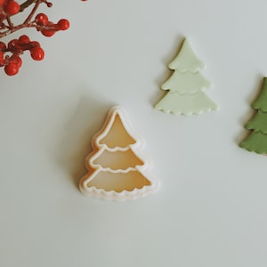 May include: Three Christmas tree cookie cutters, one white, one green and one clear plastic. The cutters are on a white surface.