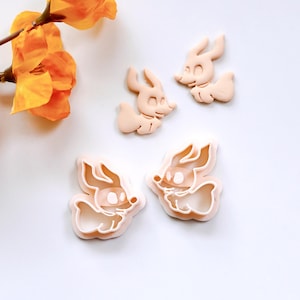 May include: Two cookie cutters in the shape of a cartoon dog with large ears and a wide smile. The cutters are made of a light pink plastic.