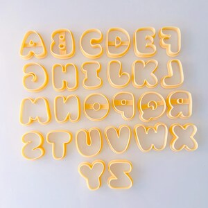 May include: A complete set of twenty-six yellow plastic alphabet cookie cutters, arranged in rows from A to Z. Each letter features a rounded, bubble-style font with internal support bars for letters like A, B, D, O, P, Q, and R to maintain their shape.