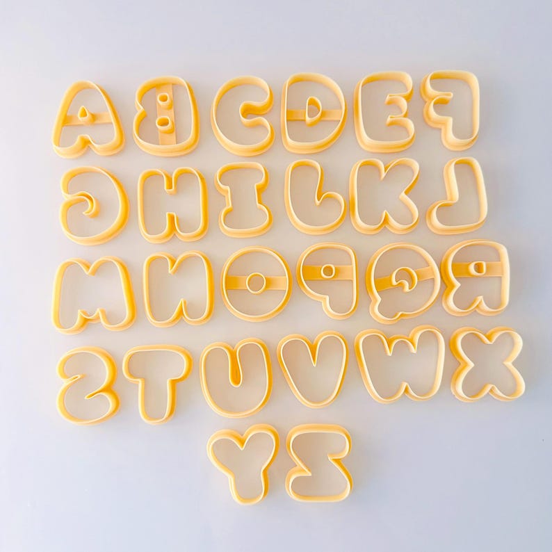 May include: A complete set of twenty-six yellow plastic alphabet cookie cutters, arranged in rows from A to Z. Each letter features a rounded, bubble-style font with internal support bars for letters like A, B, D, O, P, Q, and R to maintain their shape.