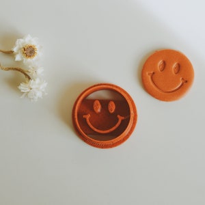May include: Two brown plastic cookie cutters in the shape of a smiley face. The cutter on the left is closed and the cutter on the right is open.