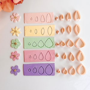 May include: Five sets of pink, yellow, green, pink, and purple fondant cutters. Each set includes three different sizes of flower petals and a small circle cutter. The cutters are arranged in rows on a white surface.