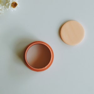Puede incluir: Un recipiente redondo de color terracota con una tapa. La tapa es de color melocotón claro y está situada junto al recipiente.