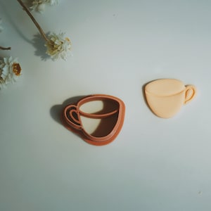 Pode incluir: Dois cortadores de biscoitos em forma de xícara de café. Um é de cor marrom claro e o outro de cor marrom escuro.