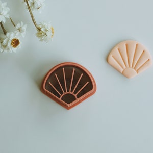 May include: A cookie cutter and a cookie in the shape of a sunburst fan. The cookie cutter is a dark brown color with a light pink interior. The cookie is light pink and has a sunburst design.