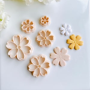 May include: Set of seven plastic flower-shaped cookie cutters in various sizes. The cutters are in shades of pink and white. There are also two fondant flowers in pink and yellow.