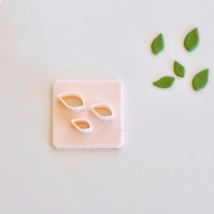 May include: A pink plastic mold with three leaf-shaped cutouts. There are five green leaf-shaped fondant pieces on a white surface.