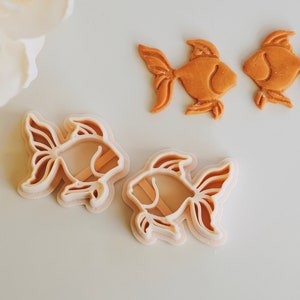 May include: Goldfish-shaped cookie cutters and baked biscuits. The cutters are light brown and have a layered design. The baked biscuits are a darker shade of brown. The image is taken on a white surface with a soft, neutral background.