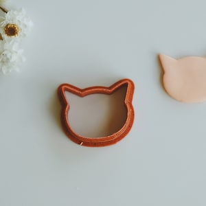 May include: A red plastic cat-shaped cookie cutter on a white surface.