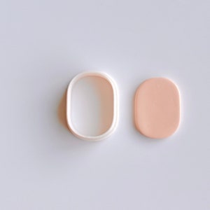 Puede incluir: Un cortador de galletas ovalado de plástico blanco y un óvalo de fondant de color melocotón claro.