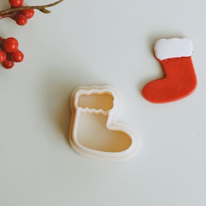 Puede incluir: Dos cortadores de galletas de calcetines de Navidad, uno blanco y otro rojo. El cortador blanco es un contorno detallado de un calcetín con una parte superior esponjosa. El cortador rojo es un calcetín sólido con una parte superior esponjosa blanca.