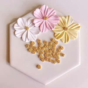 May include: A white hexagonal tile with three paper flowers in pink, white, and yellow. The flowers are arranged in a cluster in the center of the tile. Gold glitter is scattered around the flowers.
