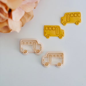 Op de afbeelding: Twee gele koekjesvormen in de vorm van schoolbussen en twee witte plastic koekjesvormen in de vorm van schoolbussen. De koekjesvormen liggen op een wit oppervlak.