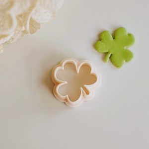 May include: A white plastic four-leaf clover cookie cutter. The cutter is on a white surface next to a green four-leaf clover.