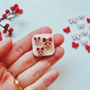 Puede incluir: Una mano sostiene un cortador de galletas cuadrado, blanquecino, con tres formas de lazo rojo. Dispersas, pequeñas decoraciones en forma de lazo rojo y blanco. Una ramita de bayas rojas en el fondo.