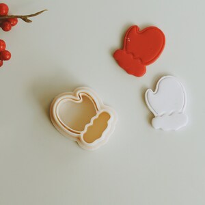 May include: Three cookie cutters shaped like mittens. One is red, one is white, and one is a clear plastic cutter.