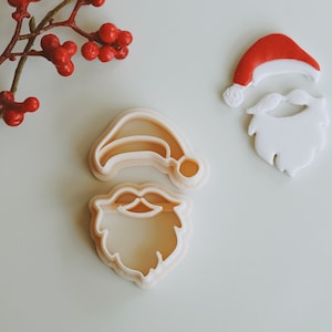 May include: Two white plastic cookie cutters shaped like Santa Claus. The top cutter is a Santa hat and the bottom cutter is a Santa beard and mustache.