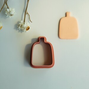 May include: Two cookie cutters in the shape of a bottle. The cutter on the left is brown and the cutter on the right is light orange.