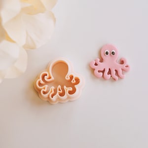 May include: A pink fondant octopus with black googly eyes, next to a plastic cookie cutter in the shape of an octopus.