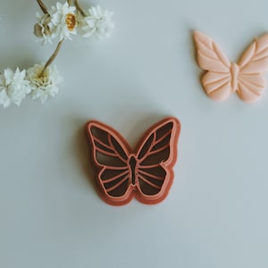 May include: Two butterfly cookie cutters, one pink and one red. The pink cutter is on a white surface and the red cutter is on a white surface with dried flowers.
