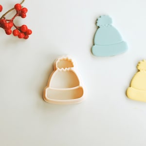 May include: Three cookie cutters shaped like winter hats. The cutters are made of white plastic and have a textured surface. One cutter is light blue, one is yellow, and one is the plain white cutter.