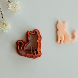 May include: Two cookie cutters in the shape of a cat. The cutter on the left is red and has a detailed design. The cutter on the right is light pink and has a simple design.