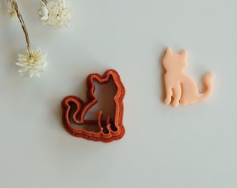 May include: Two cookie cutters in the shape of a cat. The cutter on the left is red and has a detailed design. The cutter on the right is light pink and has a simple design.