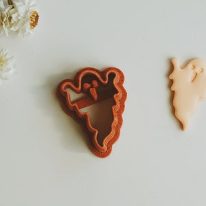 May include: A red plastic ghost cookie cutter with a white ghost cookie next to it.