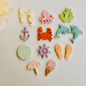 May include: A set of 12 colourful fondant toppers featuring various summer and beach themes, including an anchor, a crab, a beach ball, a starfish, a seashell, a seahorse, an octopus, a dolphin, flip flops, an ice cream cone, and an umbrella.