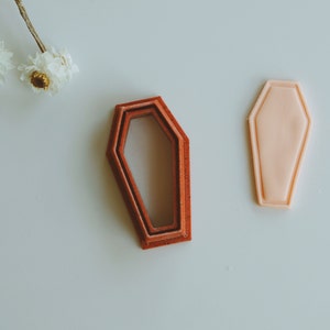 May include: Set of four nesting coffin-shaped cookie cutters. The cutters are made of red plastic and are arranged in decreasing size. The largest cutter is about 4 inches long.