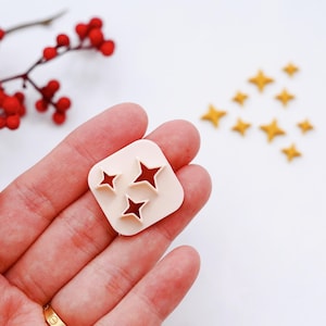 Puede incluir: Un molde de plástico blanco con tres recortes en forma de estrella. El molde está siendo sostenido en una mano. Hay varias estrellas doradas esparcidas sobre una superficie blanca.