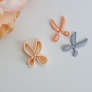 May include: Three plastic cookie cutters shaped like scissors. The cutters are in shades of light pink, peach, and gray.