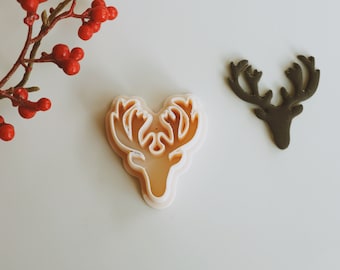 May include: Two reindeer cookie cutters, one is a light brown color and the other is a dark green color. Both cutters have a detailed design of a reindeer head with antlers.