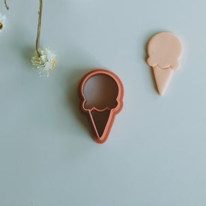 May include: Two pink plastic ice cream cone cookie cutters. One is a solid shape and the other is an outline.