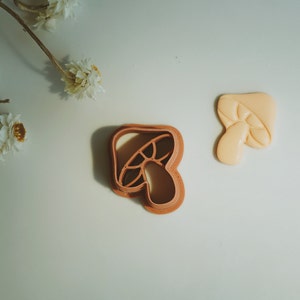 May include: Two brown plastic mushroom cookie cutters on a white surface. One cutter is closed and the other is open.