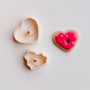 May include: Three cookie cutters in the shape of hearts. Two are plain and one has a pink donut design.