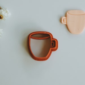 May include: A red plastic cookie cutter shaped like a coffee cup. The cutter is on a white surface.
