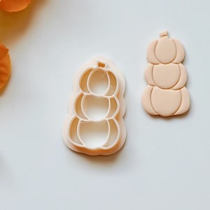 Puede incluir: Un juego de dos cortadores de galletas de plástico con forma de pila de calabazas. Los cortadores son de color melocotón claro.