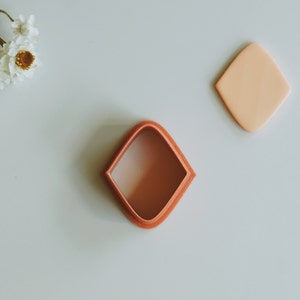 May include: A terracotta-colored diamond-shaped cookie cutter with a smooth, rounded interior. The cutter is on a white surface.