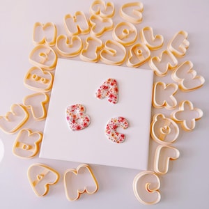 Può includere: Una collezione di formine per biscotti a forma di lettere in plastica arancione chiaro disposte attorno a una piastrella quadrata bianca. Tre lettere finite, A, B e C, sono posizionate sulla piastrella con un motivo floreale rosso.