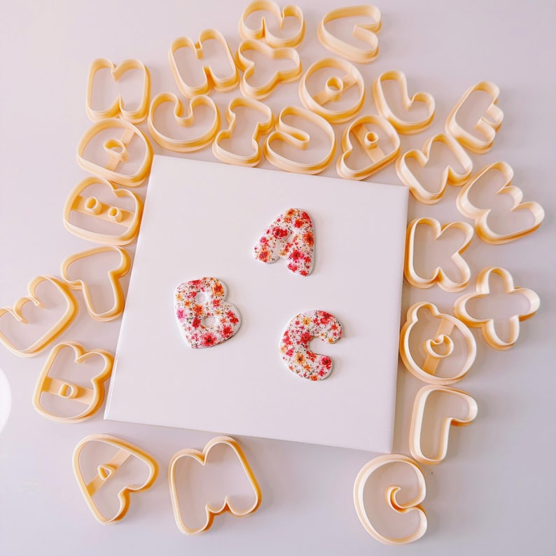 May include: A collection of light orange plastic alphabet cookie cutters arranged around a central white square tile. Three finished letter shapes, A, B, and C, are placed on the tile, each featuring a vibrant red and orange floral pattern.